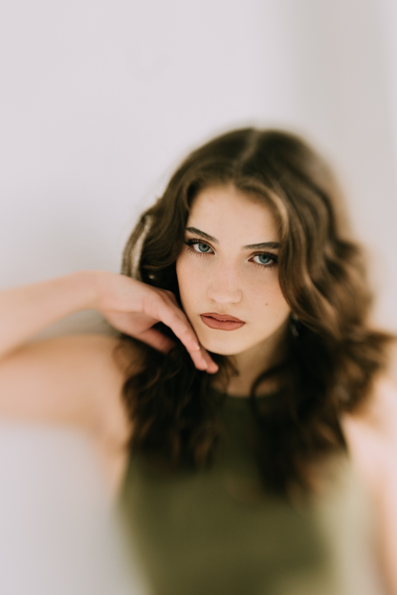 Portret młodej kobiety z brązowymi, falowanymi włosami i zielonymi oczami, pozującej z dłonią przy twarzy na jasnym tle.