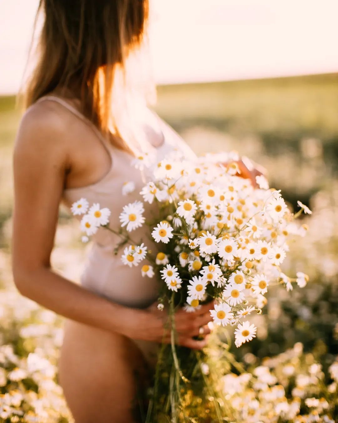 Kobieta w beżowym body trzyma duży bukiet białych margerytek na łące w złotym świetle zachodzącego słońca.