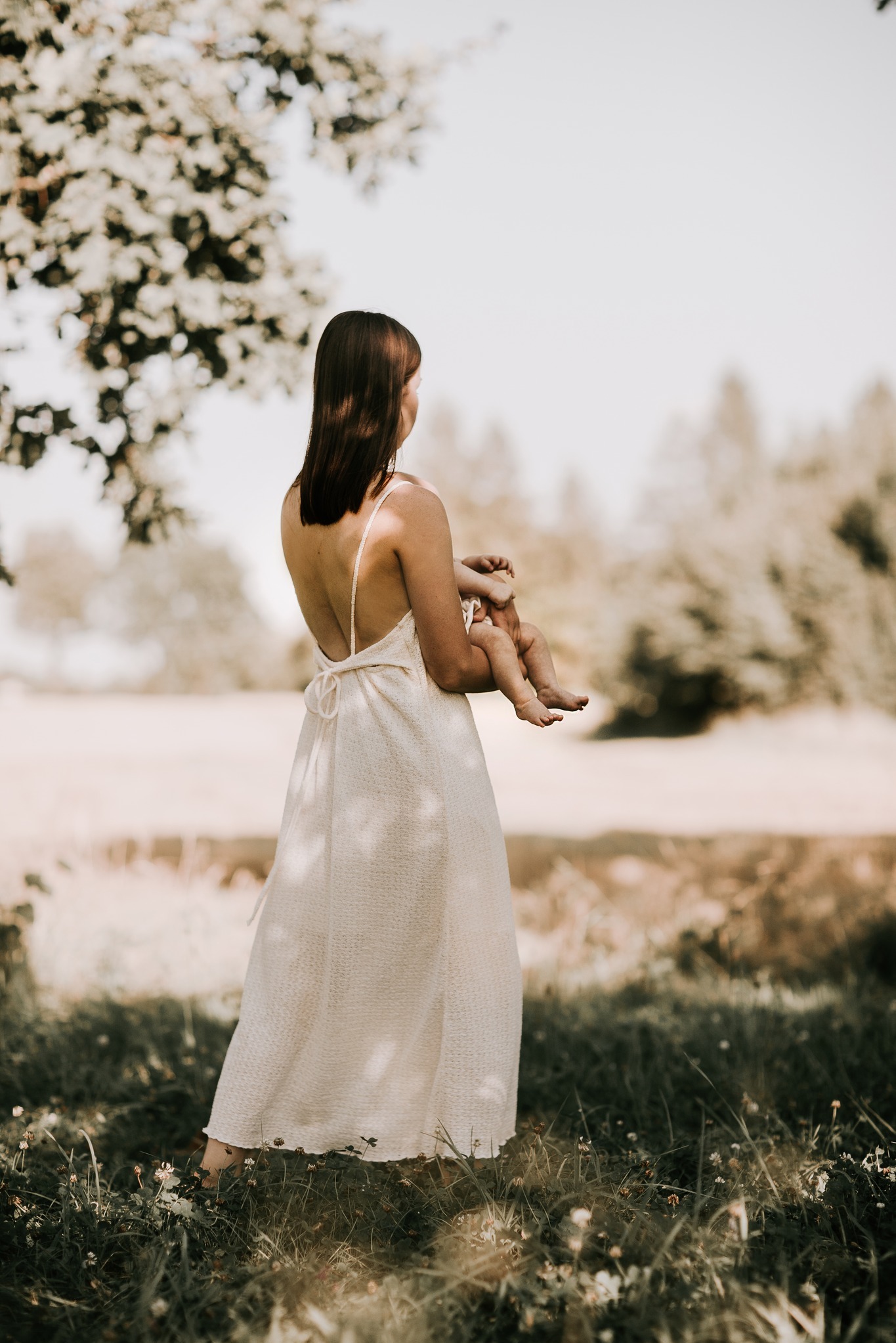 Kobieta w zwiewnej sukience z odkrytymi plecami trzymająca niemowlę na tle rozświetlonej łąki i drzew