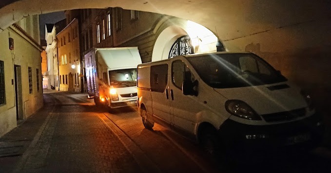 Dwa białe samochody dostawcze, jeden z zabudową kontenerową, stoją na wąskiej, oświetlonej uliczce pod arkadami w nocy.