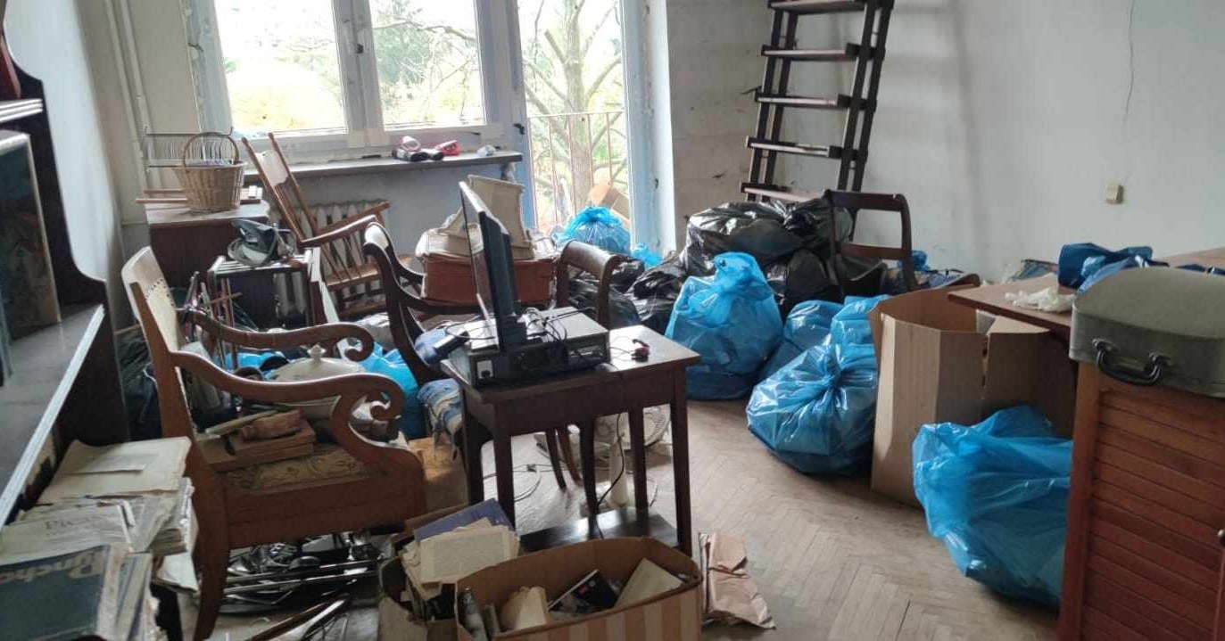 Pokój przed opróżnianiem: zagracony starymi meblami, telewizorem na stoliku, kartonami i workami na śmieci, z widoczną drabiną opartą o ścianę.