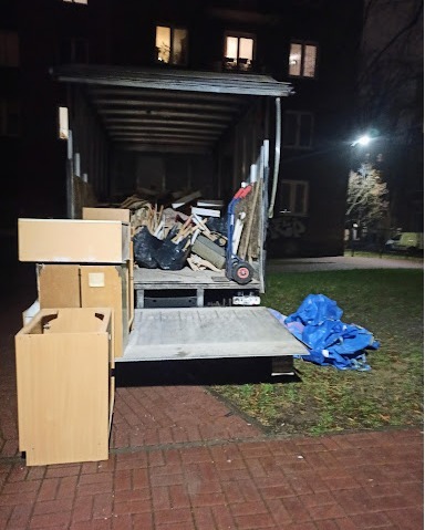 Tył ciężarówki wypełnionej meblami, kartonami i workami ze śmieciami, zaparkowanej na chodniku obok bloku mieszkalnego w nocy.