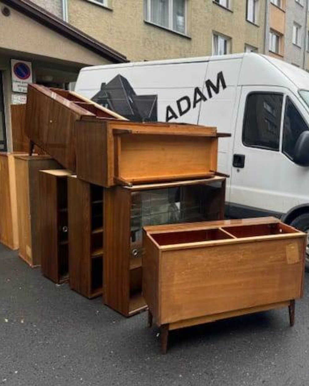 Stare, drewniane meble ustawione na zewnątrz obok białej furgonetki firmy przeprowadzkowej 'ADAM' z budynkiem mieszkalnym w tle.