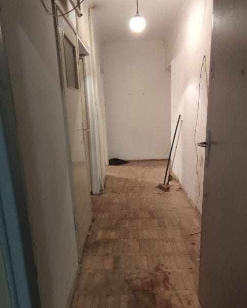 Wnętrze korytarza przed sprzątaniem, widoczne ślady zabrudzeń na podłodze z parkietu, ściany pomalowane na biało, pod sufitem wisi lampa, w tle stoi miotła.