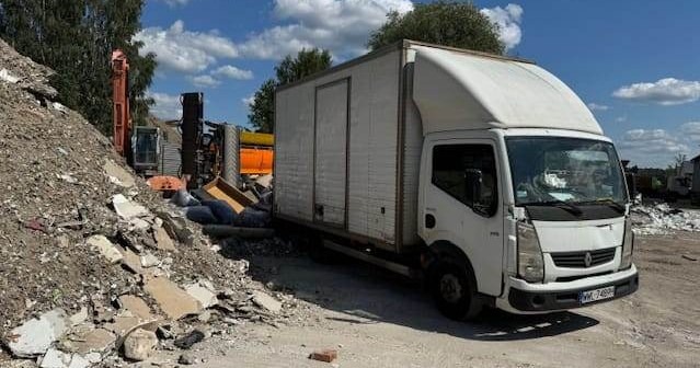 Biała furgonetka Renault zaparkowana obok sterty gruzu i wykopu z koparką w tle, prawdopodobnie podczas prac porządkowych.