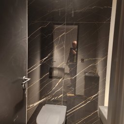 Firma-Remontowo-Budowlana Strzępek Przemysław - Wykończona łazienka z czarnymi płytkami imitującymi marmur z złotymi żyłami, wisząca toaleta w kolorze szarym, widoczne odbicie osoby robiącej zdjęcie w lustrze.