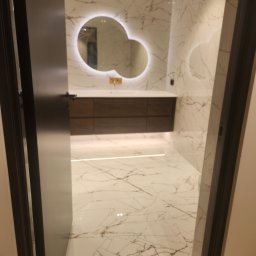 Firma-Remontowo-Budowlana Strzępek Przemysław - Nowoczesna łazienka z marmurowymi płytkami na ścianach i podłodze, okrągłe podświetlane lustro nad drewnianą szafką z umywalką, widok przez otwarte drzwi.