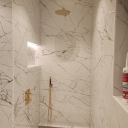 Firma-Remontowo-Budowlana Strzępek Przemysław - Kabina prysznicowa wyłożona białymi płytkami imitującymi marmur z złotą armaturą, odpływem liniowym i wnęką z podświetleniem LED, widoczna tuba silikonu na półce.