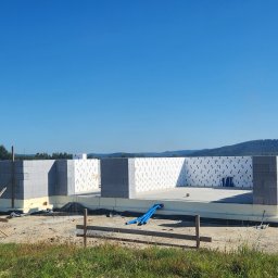 Firma-Remontowo-Budowlana Strzępek Przemysław - Wznoszenie ścian parteru domu jednorodzinnego z bloczków betonu komórkowego Ytong, widoczne izolacja fundamentów i podłoże betonowe, w tle krajobraz z zalesionymi wzgórzami.
