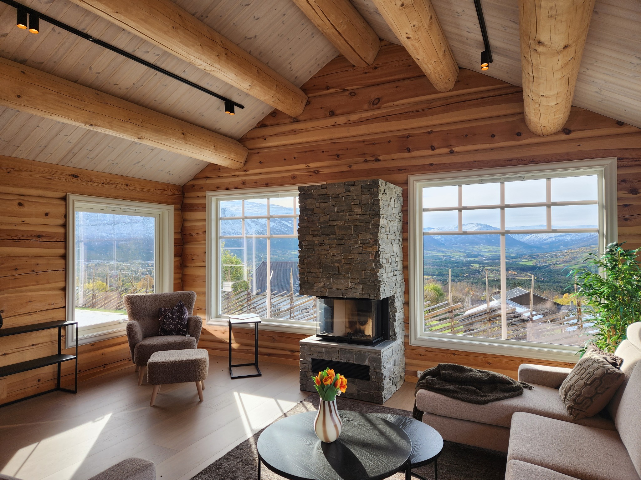 Jasne wnętrze drewnianego domu z widokiem na góry, kamienny kominek, okrągły stolik kawowy z kwiatami, fotel z podnóżkiem i sofa.