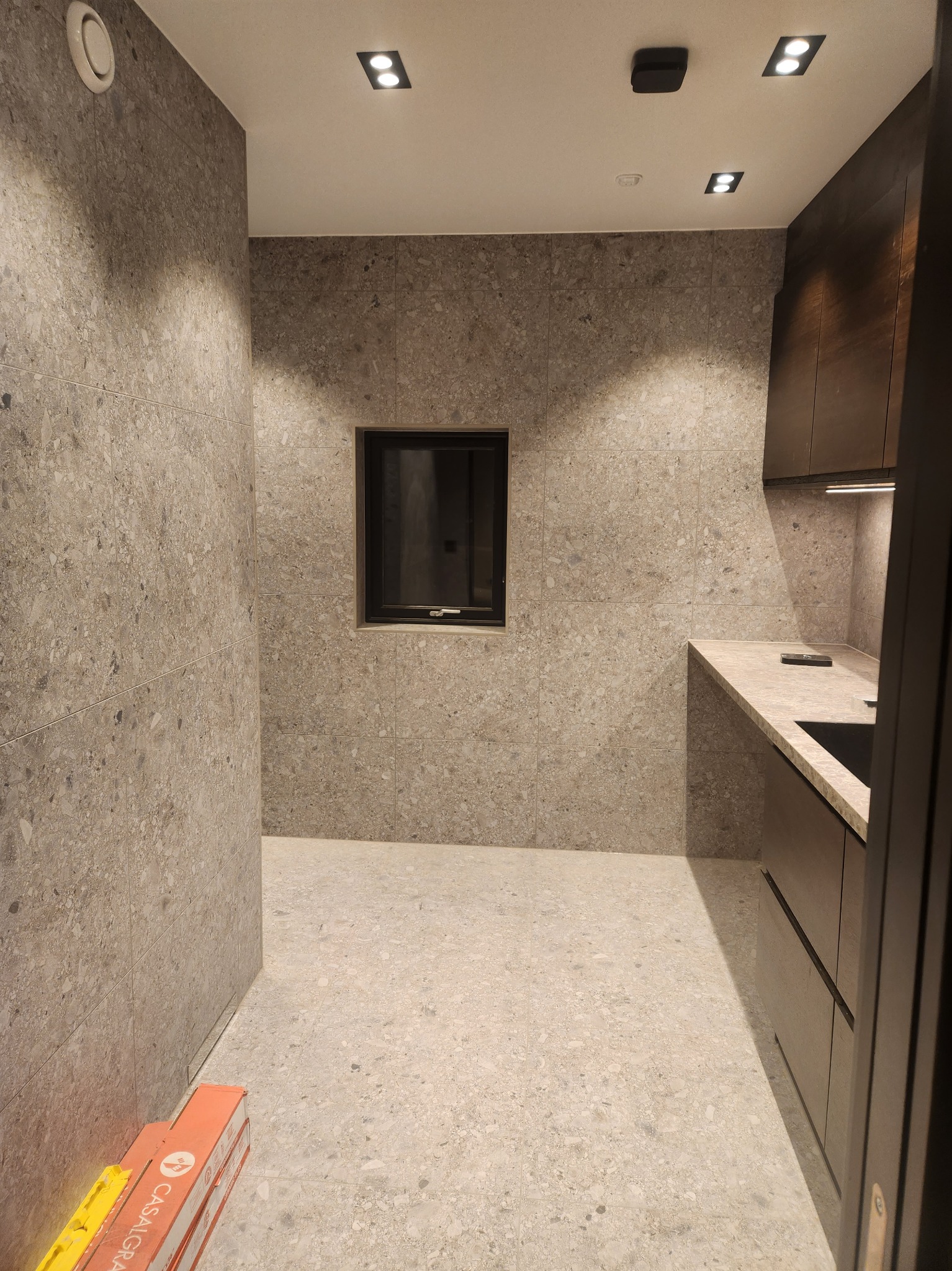 Wykończona łazienka z szarymi płytkami imitującymi lastryko na ścianach i podłodze, czarne akcenty w postaci okna i szafek, widoczne kartony z płytkami Casalgrande Padana.