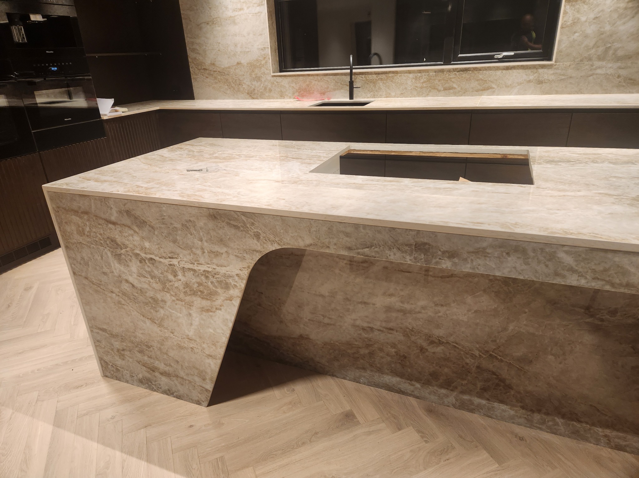 Beżowa marmurowa wyspa kuchenna z wyciętym otworem na zlew, z zabudowaną kuchenką Miele w tle, parkiet w jodełkę na podłodze.