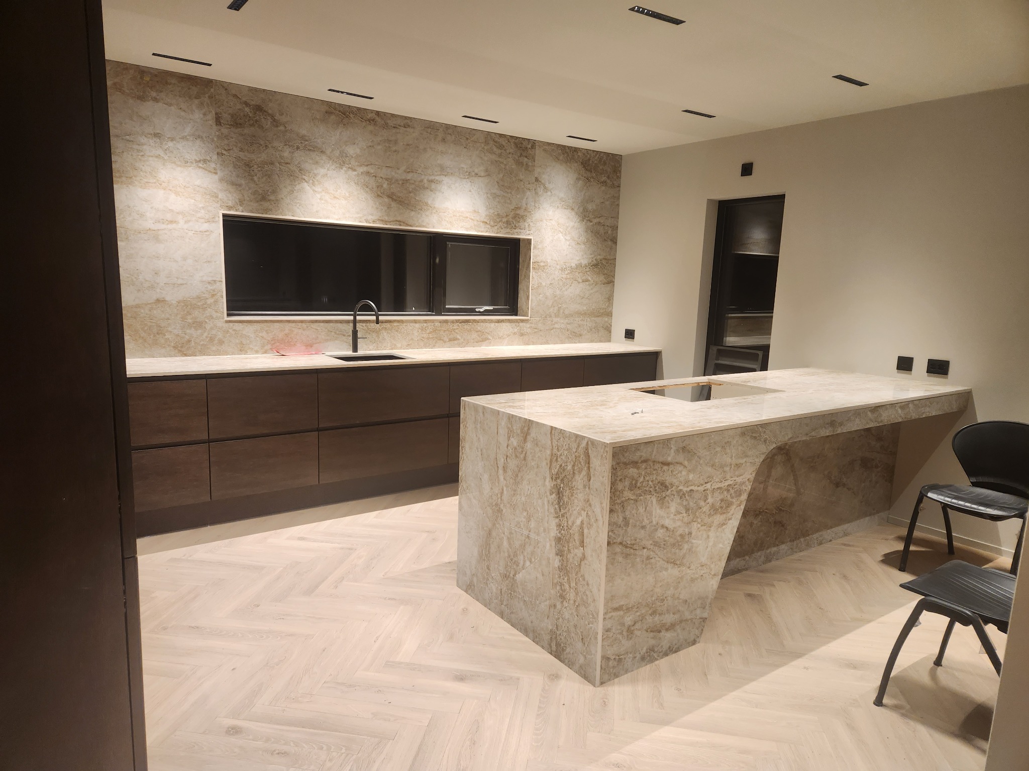 Nowoczesna kuchnia z wyspą wykończoną marmurem, ciemne szafki kuchenne pod oknem, parkiet ułożony w jodełkę.