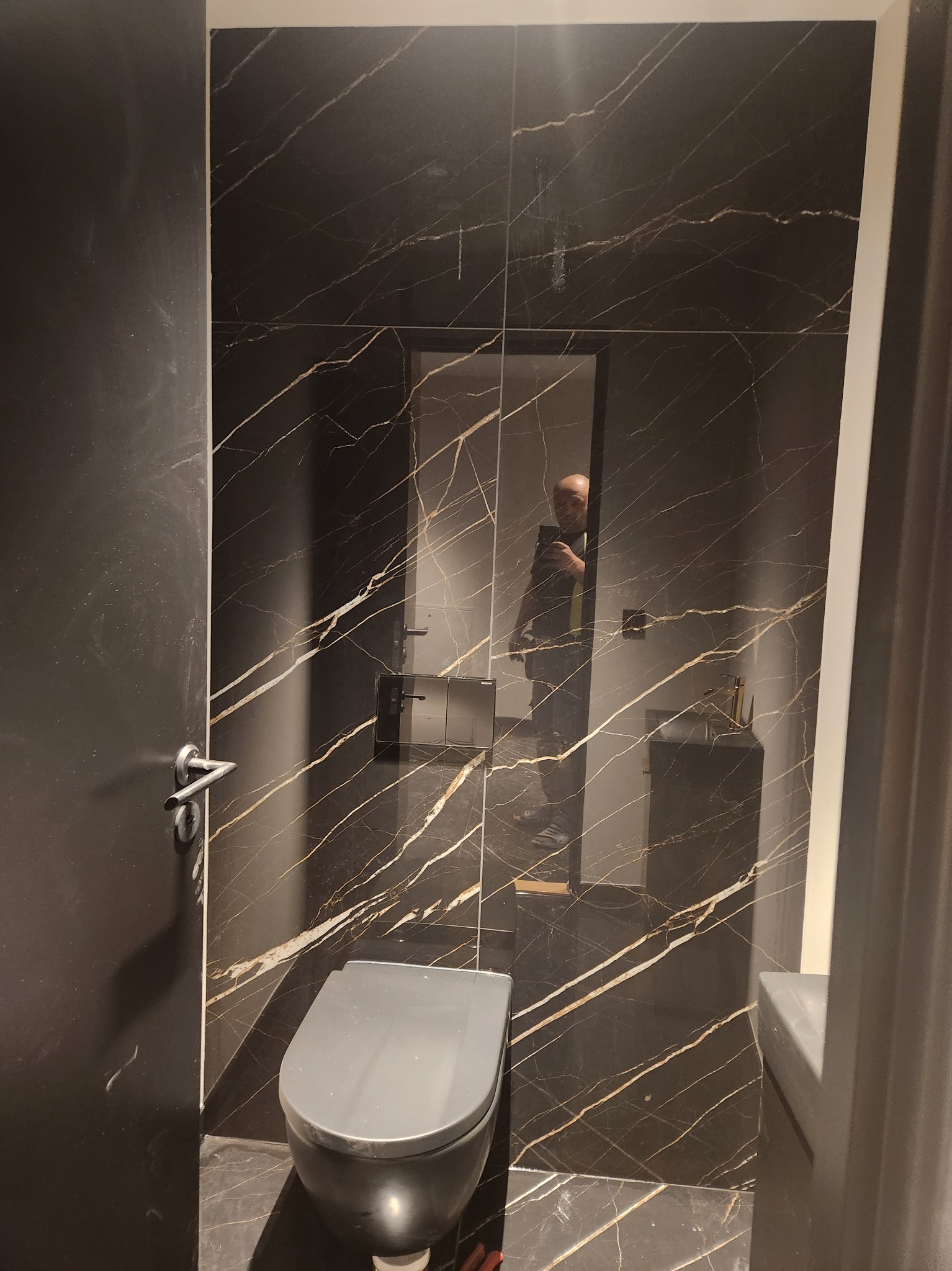 Wykończona łazienka z czarnymi płytkami imitującymi marmur z złotymi żyłami, wisząca toaleta w kolorze szarym, widoczne odbicie osoby robiącej zdjęcie w lustrze.
