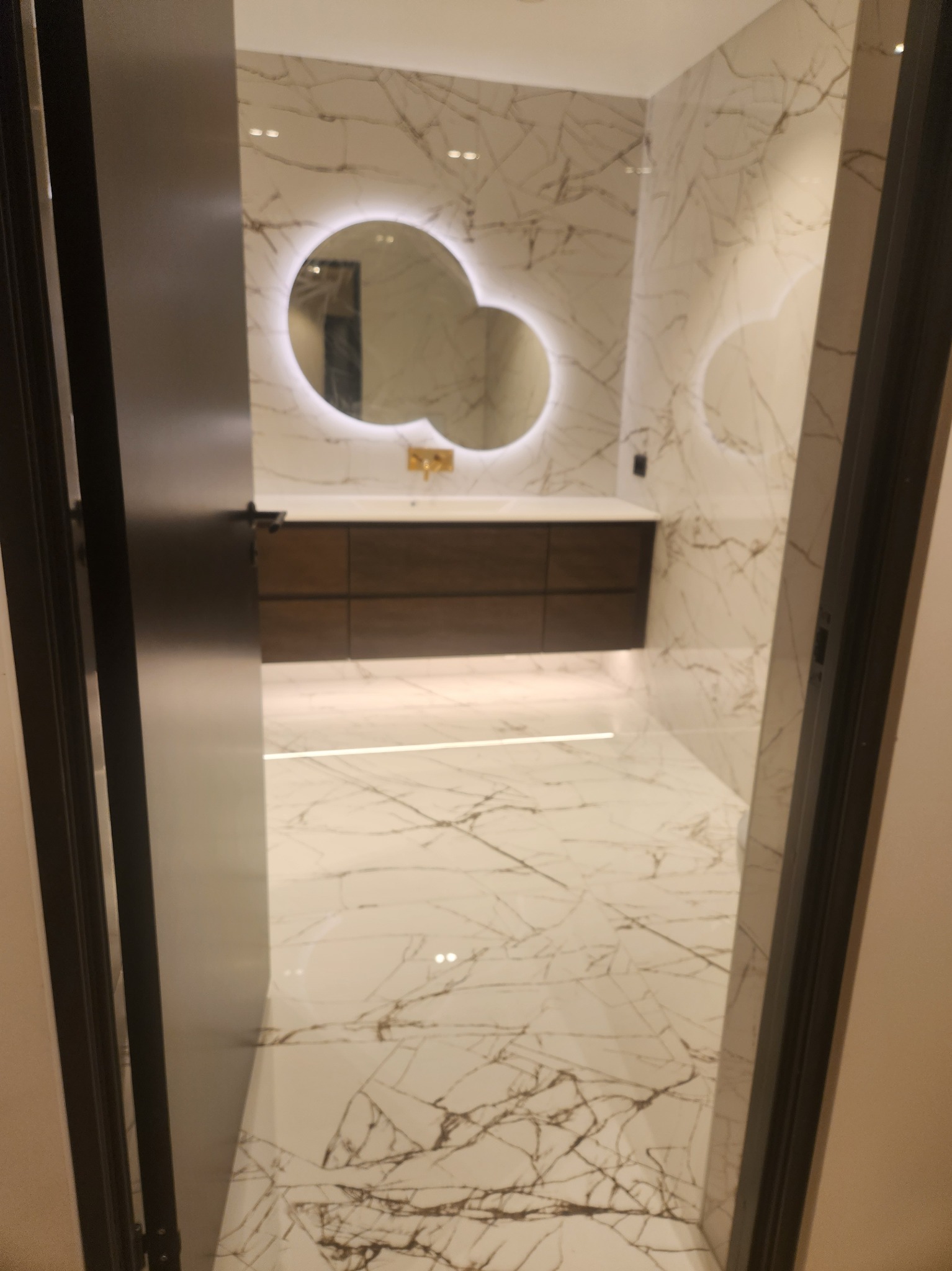 Nowoczesna łazienka z marmurowymi płytkami na ścianach i podłodze, okrągłe podświetlane lustro nad drewnianą szafką z umywalką, widok przez otwarte drzwi.