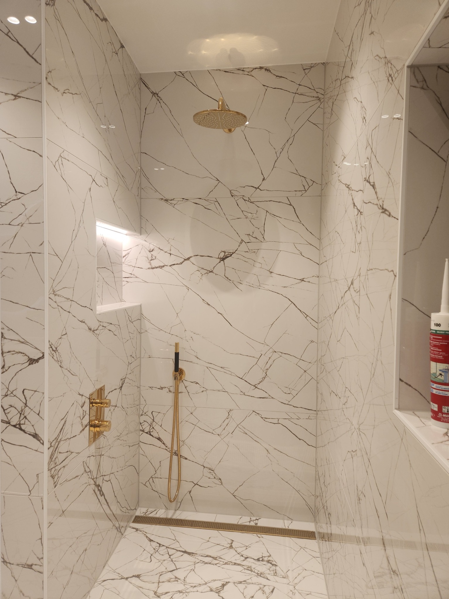 Kabina prysznicowa wyłożona białymi płytkami imitującymi marmur z złotą armaturą, odpływem liniowym i wnęką z podświetleniem LED, widoczna tuba silikonu na półce.