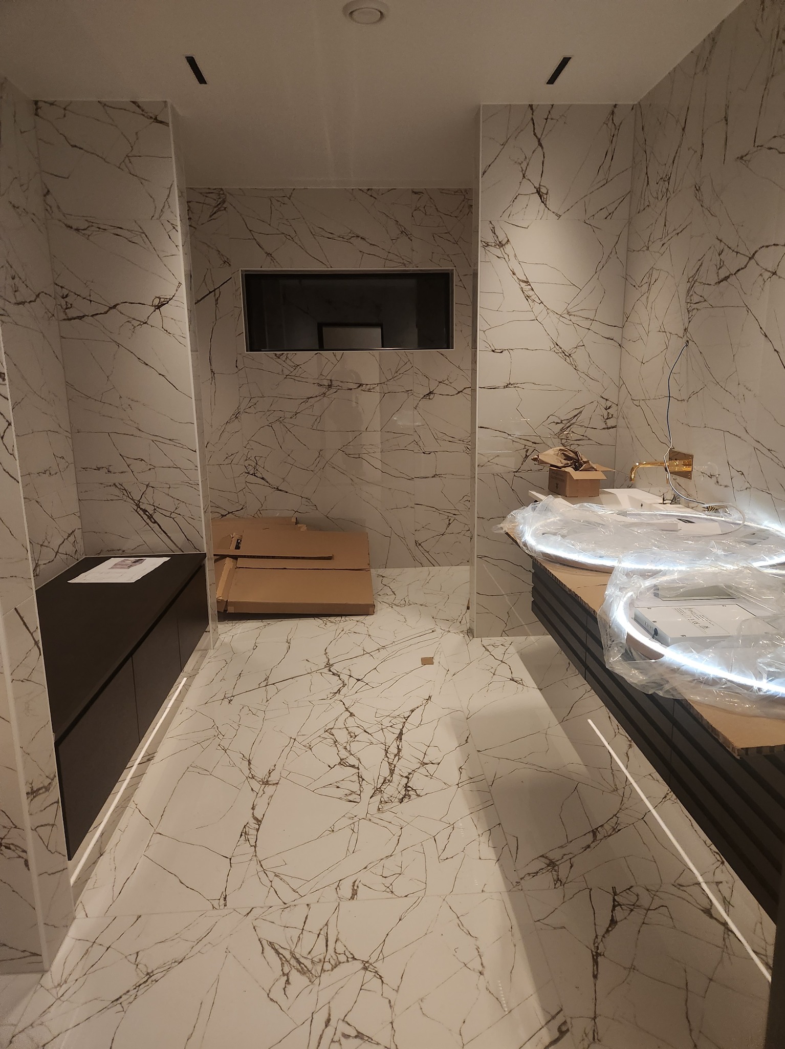 Wnętrze łazienki w trakcie wykańczania, widoczne płytki imitujące marmur na ścianach i podłodze, zabudowa szafkowa w ciemnym kolorze, złota bateria umywalkowa oraz okrągłe lustro z podświetleniem...