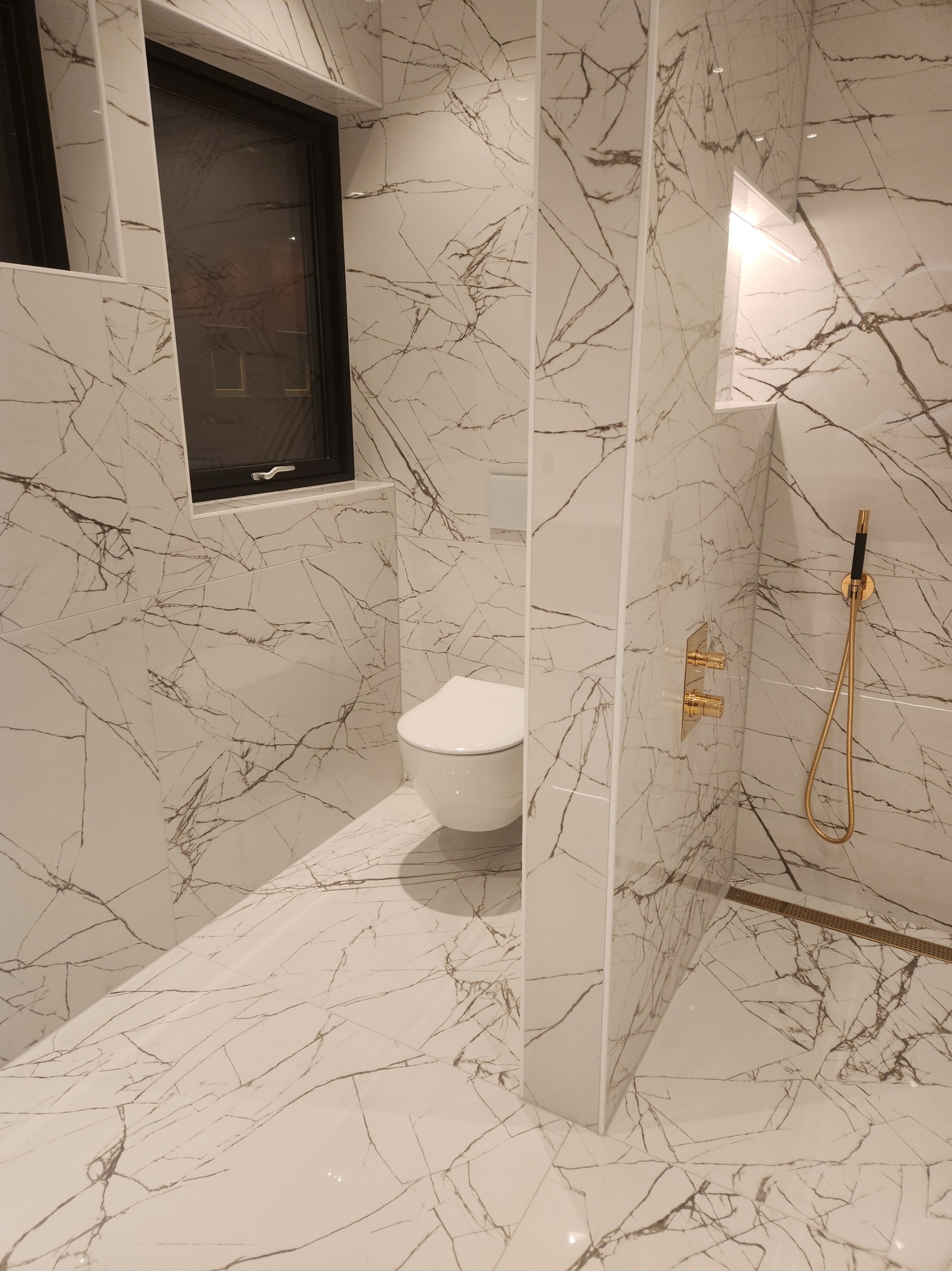 Nowoczesna łazienka wykończona białymi płytkami imitującymi marmur, z czarnym oknem, złotą armaturą i podwieszaną toaletą.