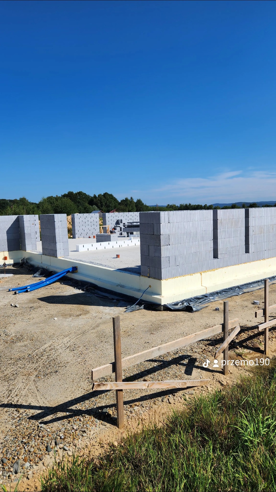 Ściany domu w trakcie budowy z szarych bloczków, widoczne niebieskie rury i ocieplenie fundamentów, tło z zielenią i błękitnym niebem.