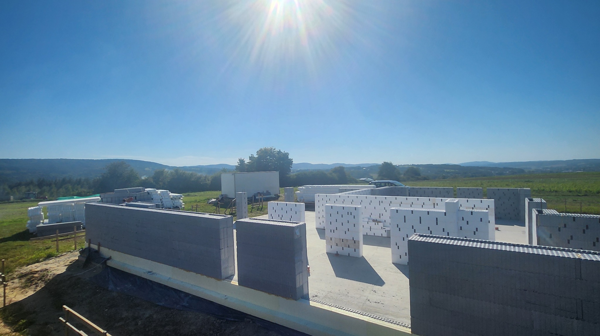 Wznoszenie ścian domu z szarego i białego betonu komórkowego na fundamencie, widok z góry na tle krajobrazu z polami i lasami w słoneczny dzień.