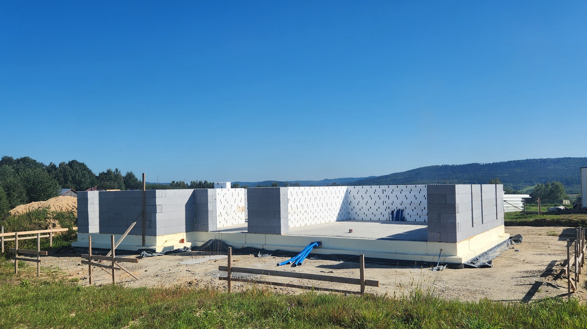 Wznoszenie ścian parteru domu jednorodzinnego z bloczków betonu komórkowego Ytong, widoczne izolacja fundamentów i podłoże betonowe, w tle krajobraz z zalesionymi wzgórzami.