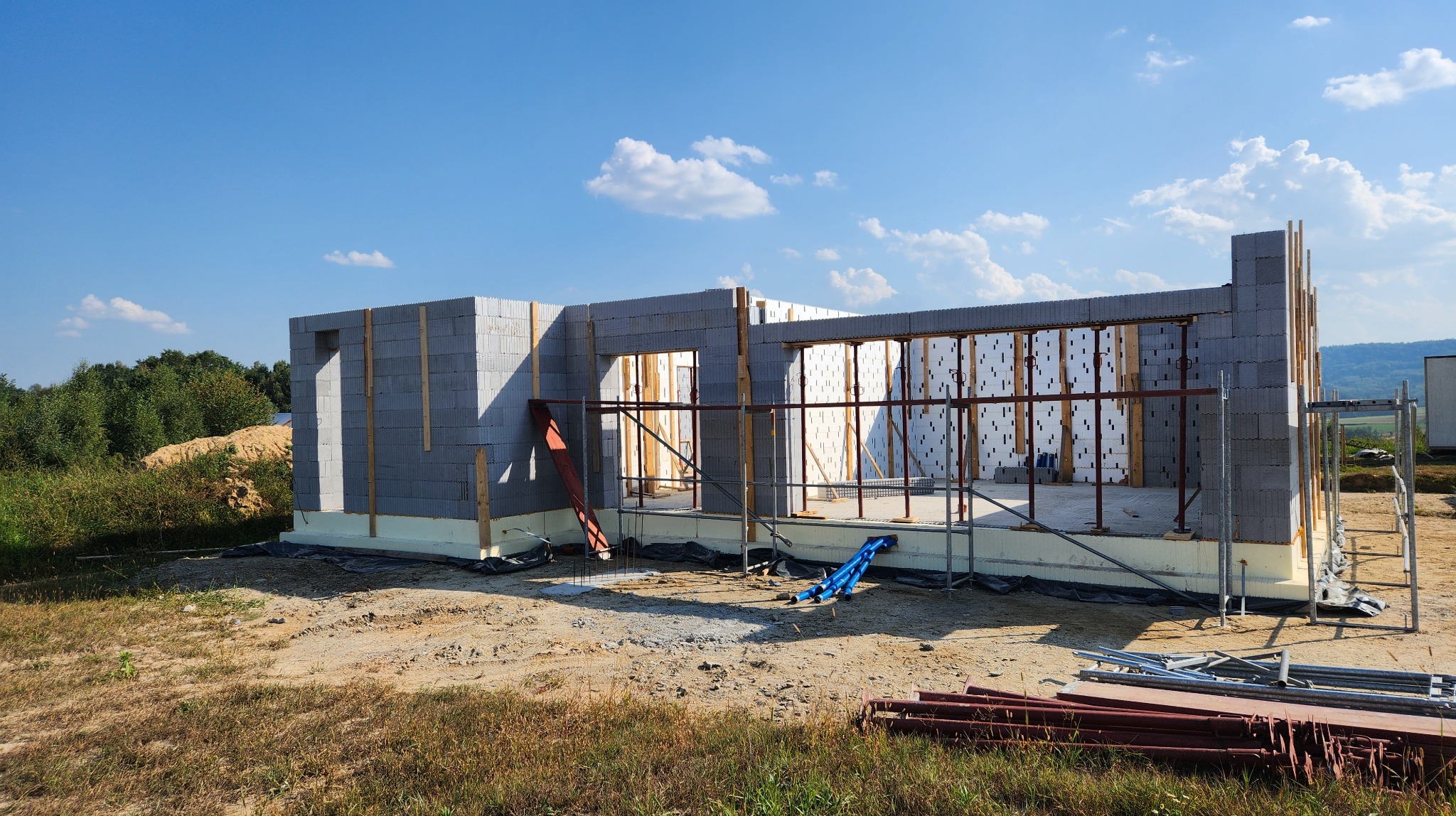 Budowa domu jednorodzinnego: ściany wzniesione z betonu komórkowego, widoczne szalunki i rusztowania, przygotowane otwory okienne, fundament ocieplony styropianem.