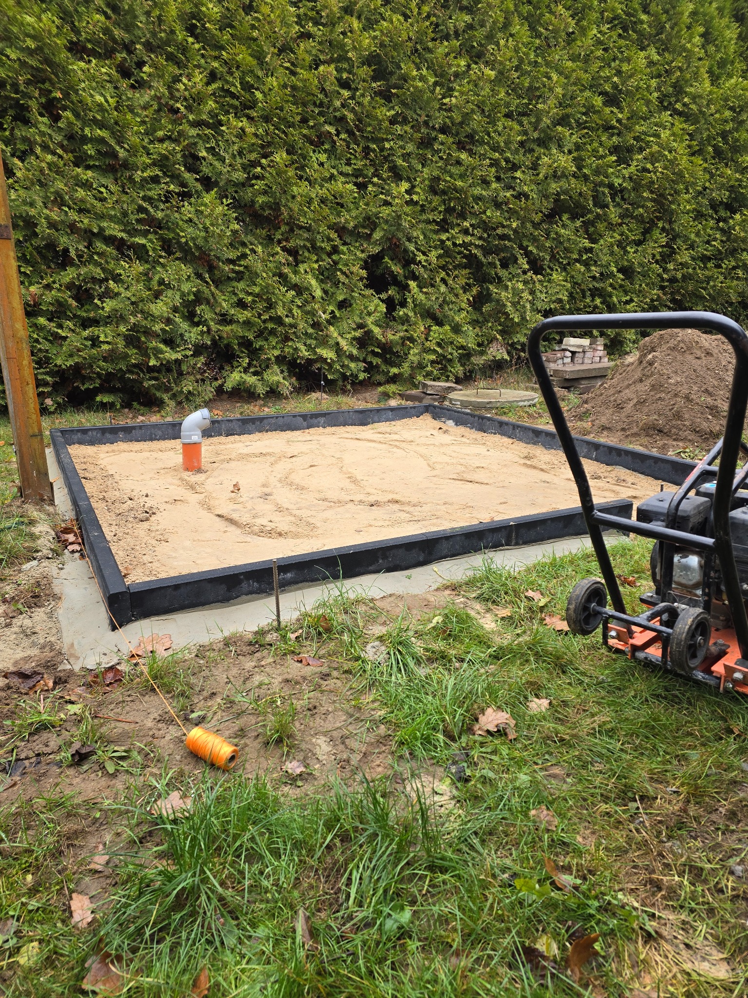 Przygotowany teren pod budowę, obramowany czarną kostką, wypełniony piaskiem, z widoczną rurą i zagęszczarką w tle, otoczony zielenią.