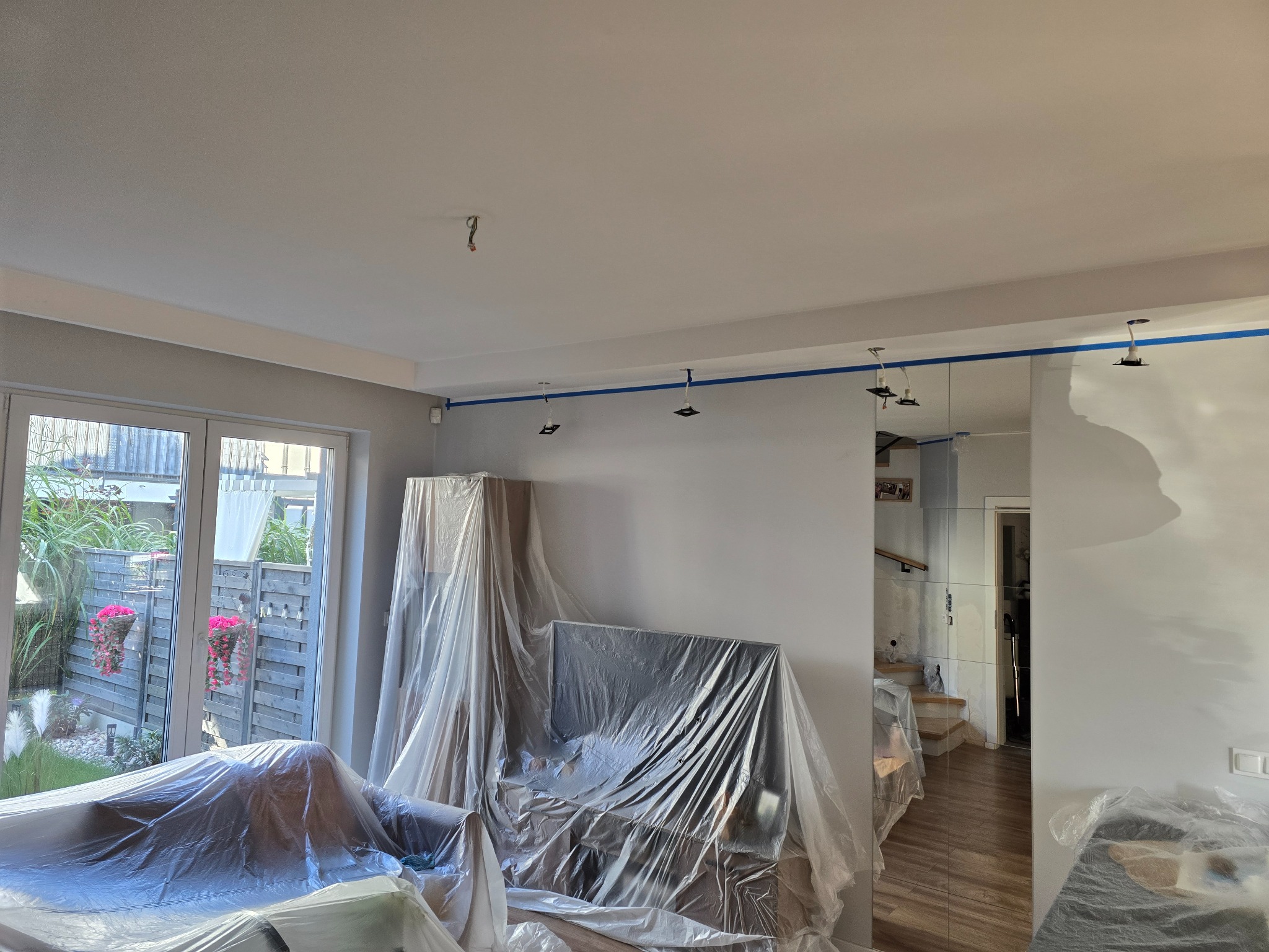 Przygotowanie pokoju do malowania: meble zabezpieczone folią, ściany z taśmą malarską, widok na okno z ogrodem i kwiatami.