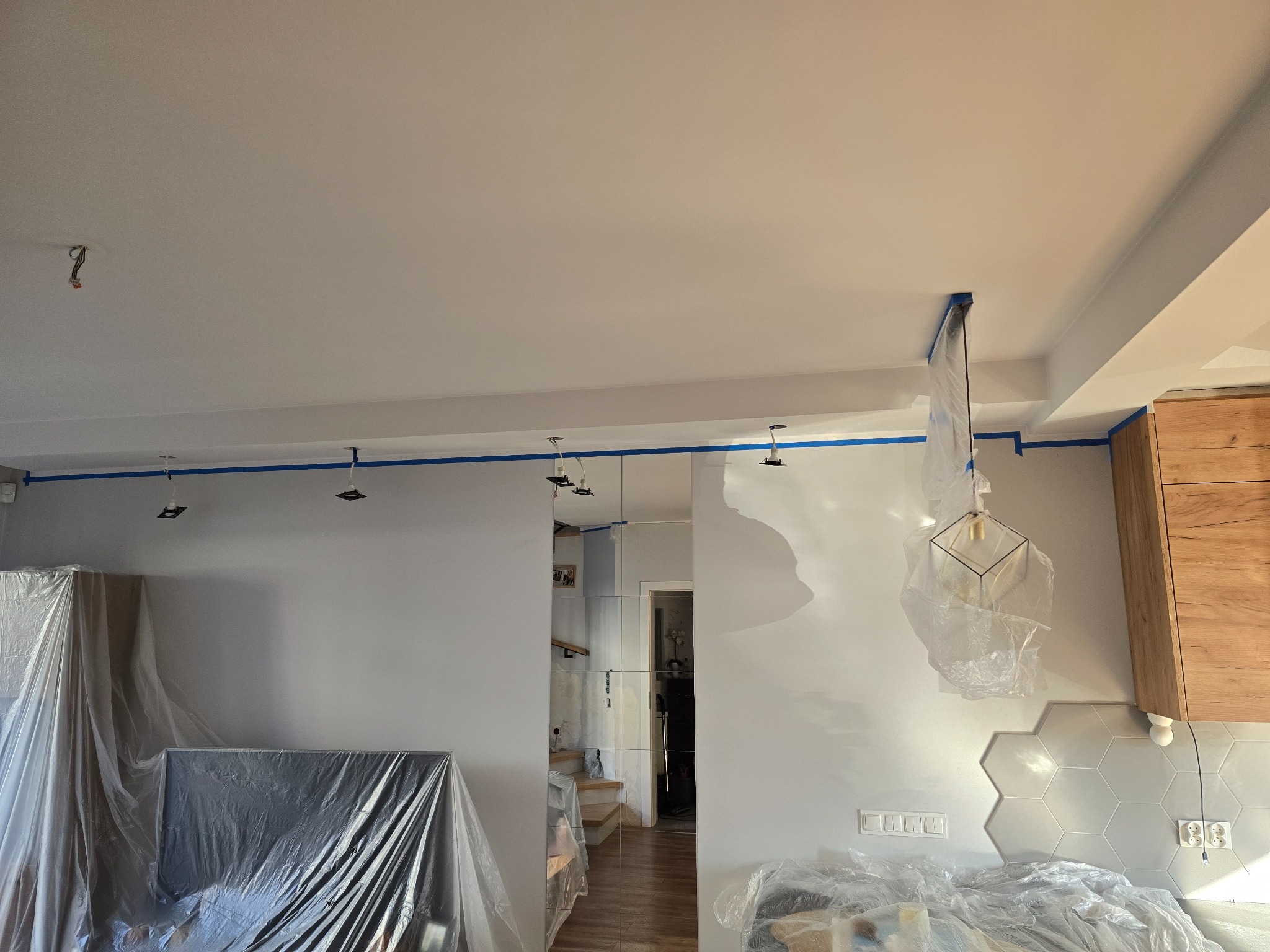 Wnętrze w trakcie malowania: ściany zabezpieczone taśmą malarską, meble przykryte folią ochronną, widoczne świeżo pomalowane ściany i sufit.