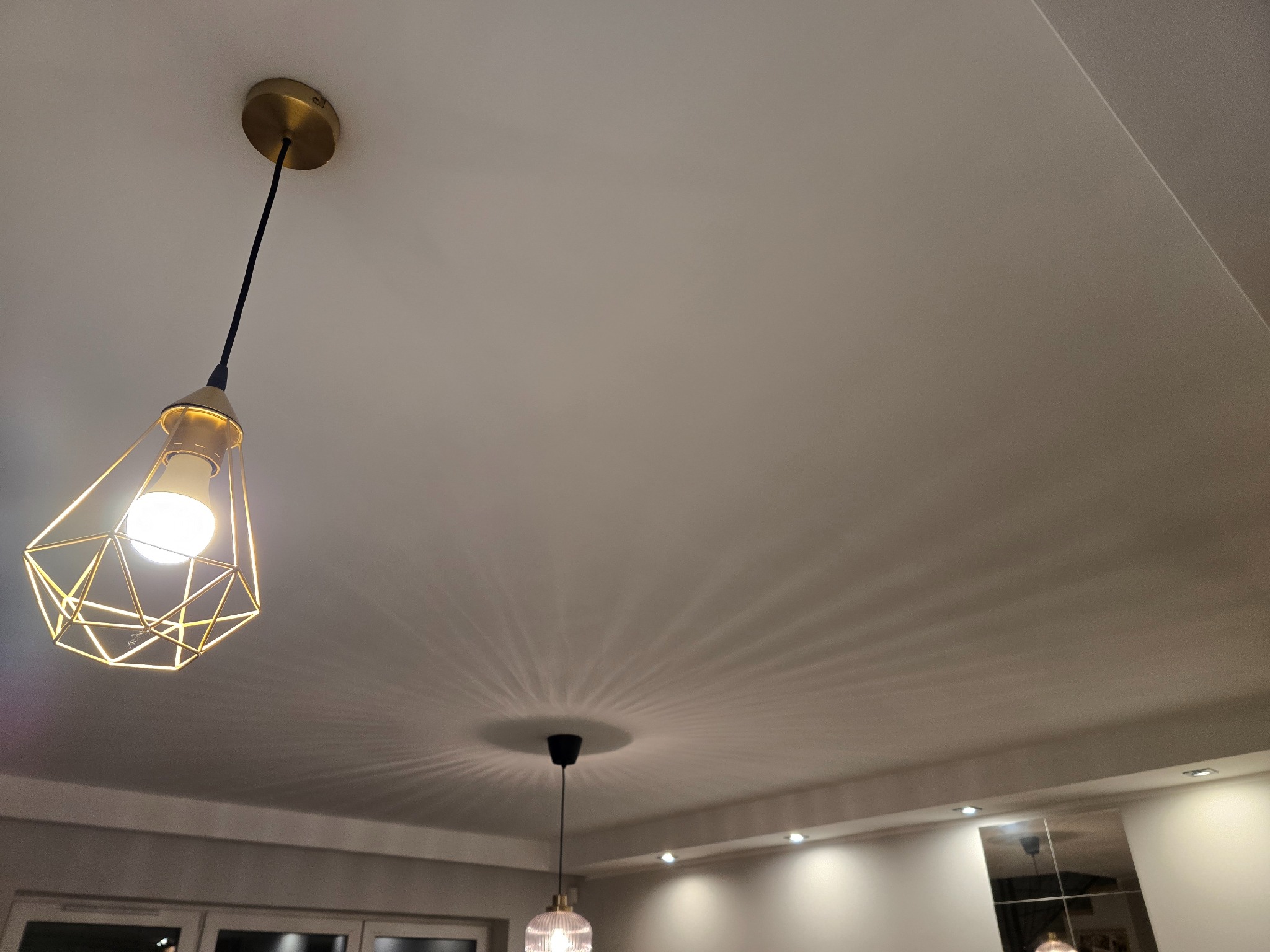 Biały sufit z dwoma lampami wiszącymi: geometryczną, złotą z lewej i szklaną, okrągłą w centrum. Widoczne światło z lamp tworzy wzory na suficie.