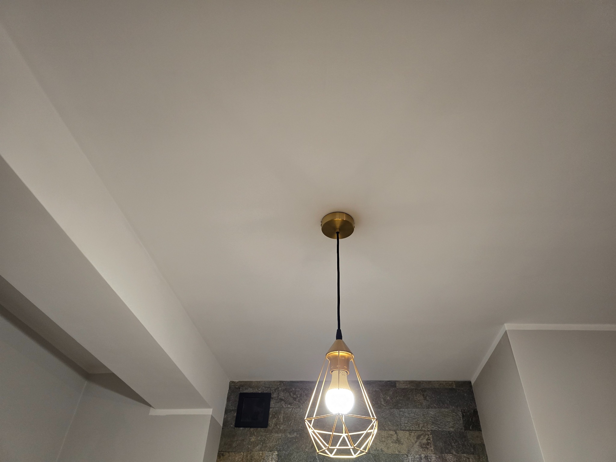 Nowoczesna lampa wisząca z geometrycznym kloszem, oświetlająca fragment białego sufitu i szarej ściany z dekoracyjnym kamieniem. Oświetlenie wnętrza.