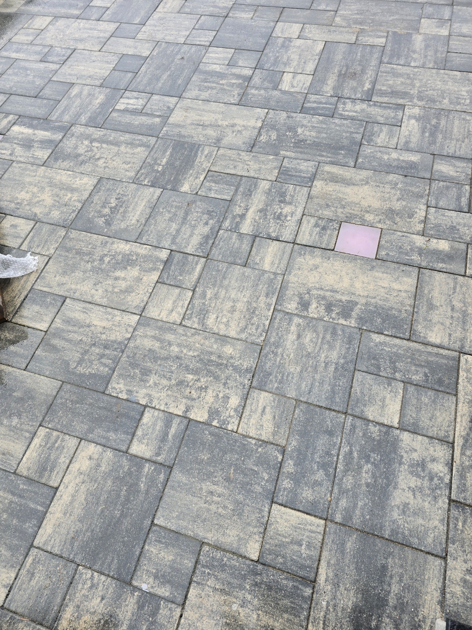Ułożona kostka brukowa w odcieniach szarości, z pojedynczym, wyróżniającym się elementem w kolorze różowym. Widoczny fragment krawężnika.