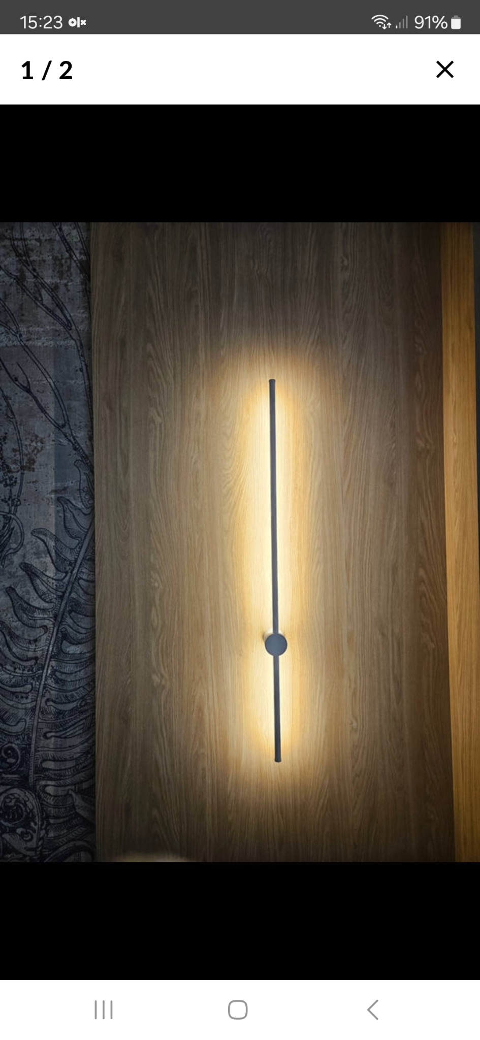 Nowoczesna lampa ścienna LED, pionowa, z okrągłym elementem pośrodku, oświetlająca drewnianą ścianę z subtelnym wzorem, tworząca ciepłą atmosferę.