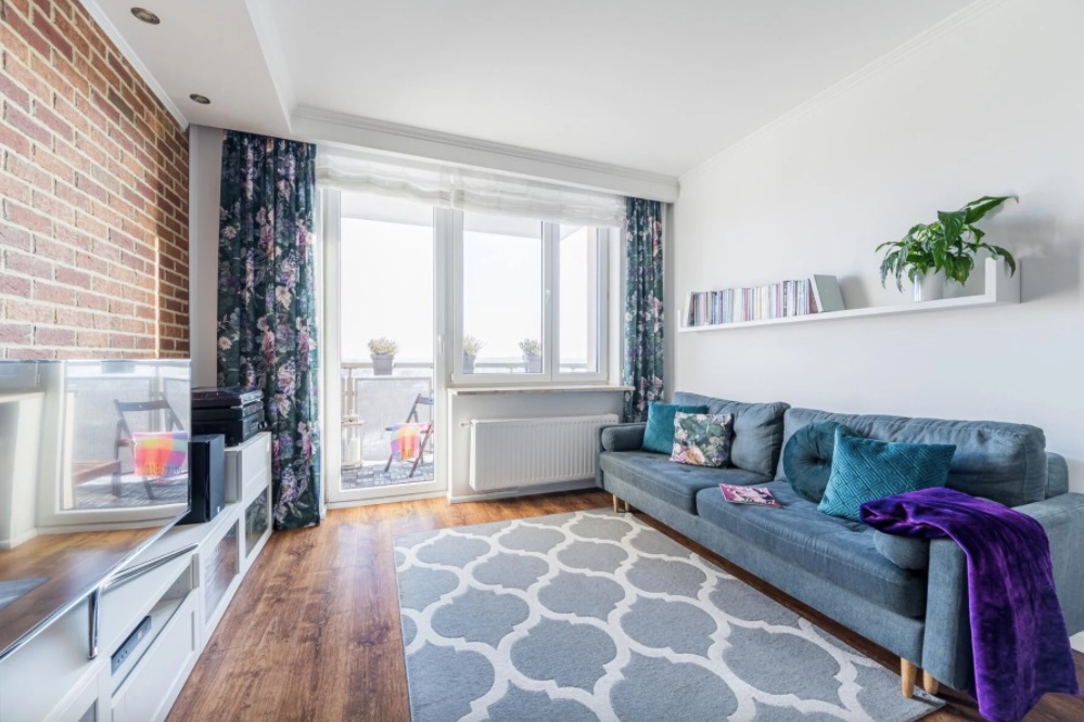 Jasny salon z ceglaną ścianą, szarą sofą z turkusowymi poduszkami i balkonem z kwiatami.