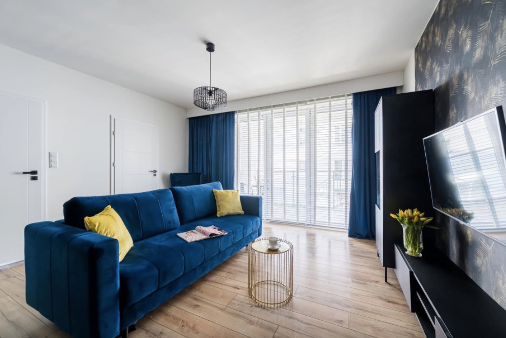 Nowoczesny salon z granatową sofą, żółtymi poduszkami, złocistym stolikiem kawowym i telewizorem na czarnej szafce RTV z bukietem żółtych tulipanów.