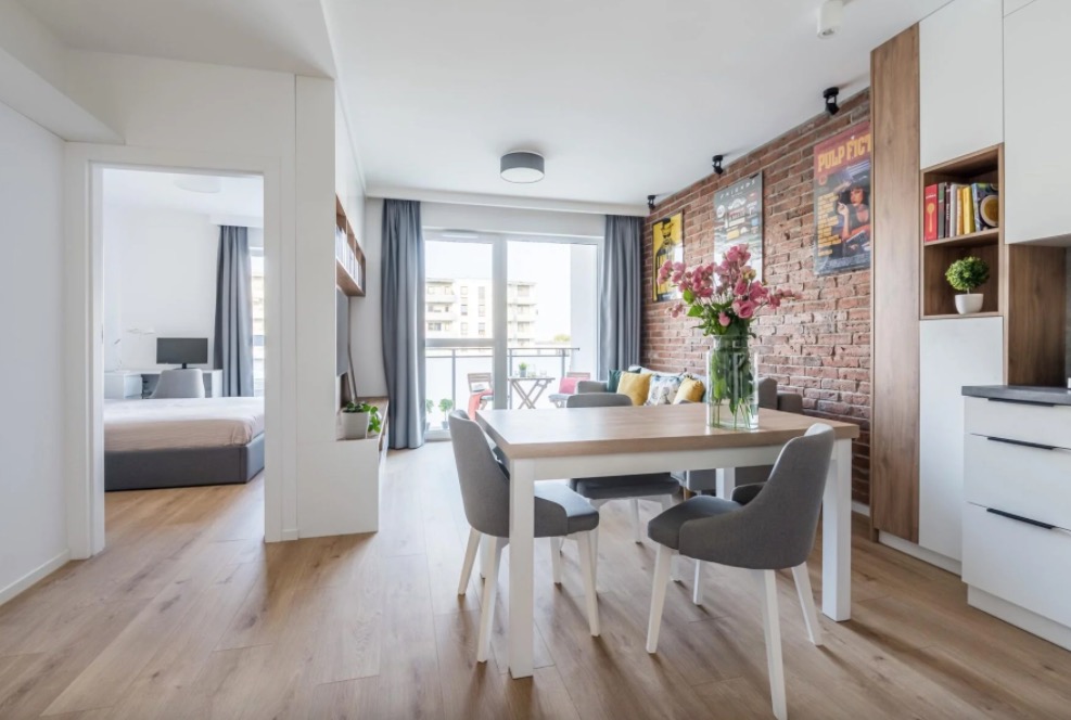 Jasne, nowoczesne wnętrze mieszkania z drewnianą podłogą, ceglaną ścianą, stołem jadalnym z szarymi krzesłami i widokiem na balkon oraz sypialnię w tle.