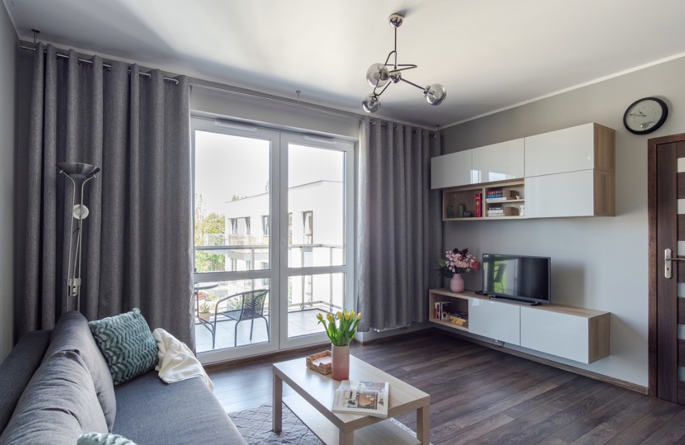 Jasne wnętrze salonu z szarą sofą, drewnianą podłogą i balkonem z widokiem na budynek mieszkalny, oświetlone nowoczesną lampą wiszącą i ozdobione kwiatami.