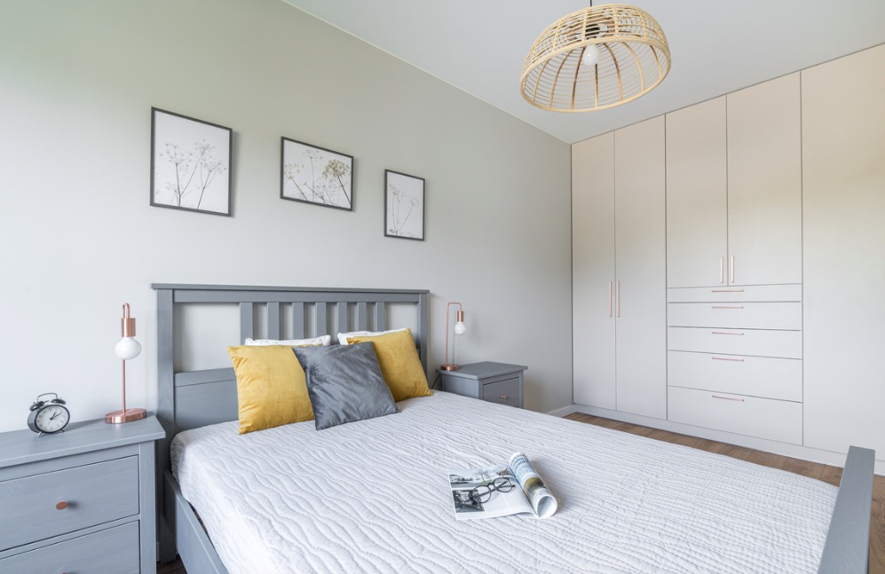 Jasna sypialnia w szarościach i żółtych akcentach: łóżko z szarą ramą i żółtymi poduszkami, szare szafki nocne z lampkami i zegarem, na ścianie grafiki roślinne w czarnych ramach, duża szafa...