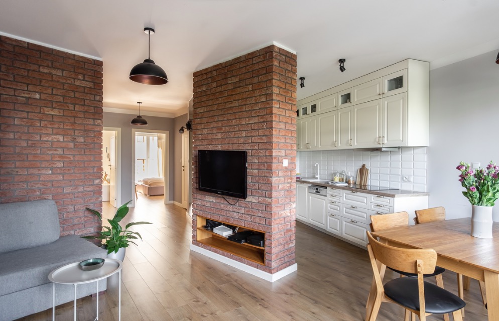 Nowoczesne wnętrze mieszkania z ceglaną ścianą działową, telewizorem, drewnianym stołem i białą kuchnią w stylu retro.