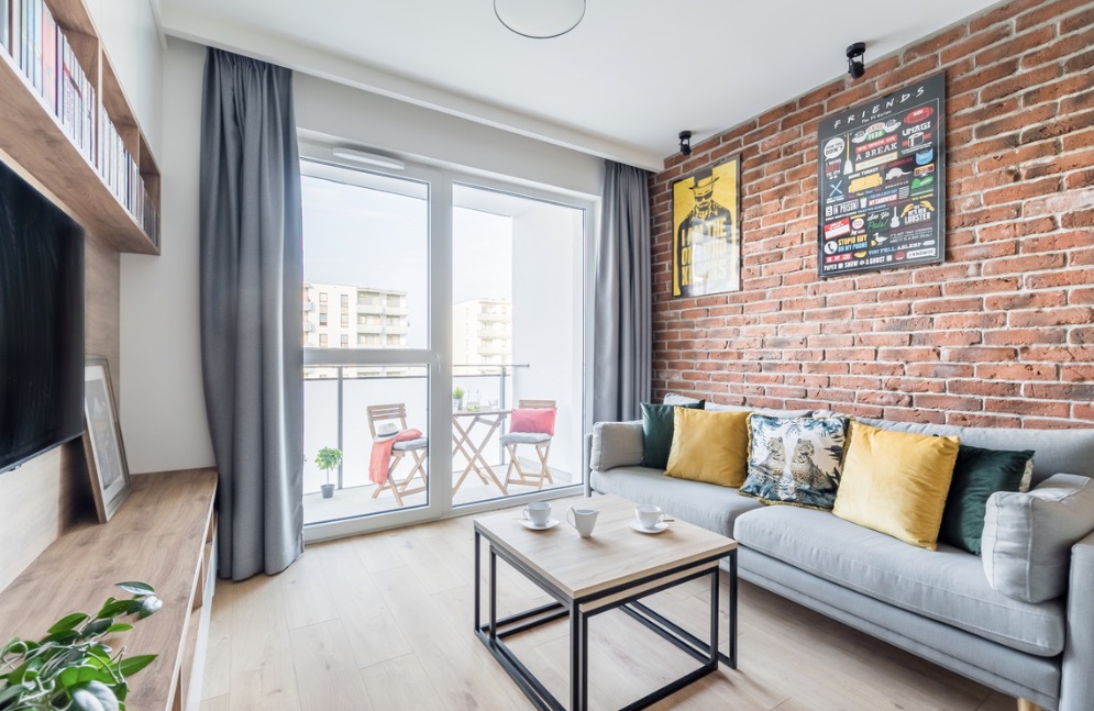 Jasne wnętrze salonu w Warszawie z ceglaną ścianą, szarą sofą z kolorowymi poduszkami, stolikiem kawowym z dwoma filiżankami i balkonem z meblami ogrodowymi.