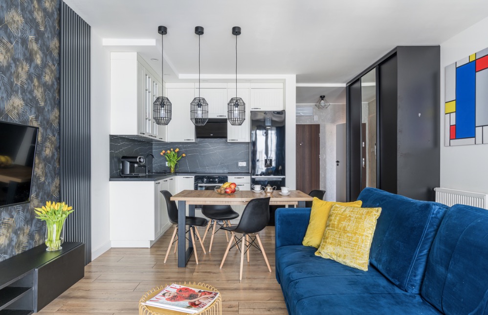Jasne wnętrze mieszkania z otwartą kuchnią, drewnianym stołem jadalnym i niebieską sofą z żółtymi poduszkami, dekoracje w postaci kwiatów i obrazu w stylu Mondriana.