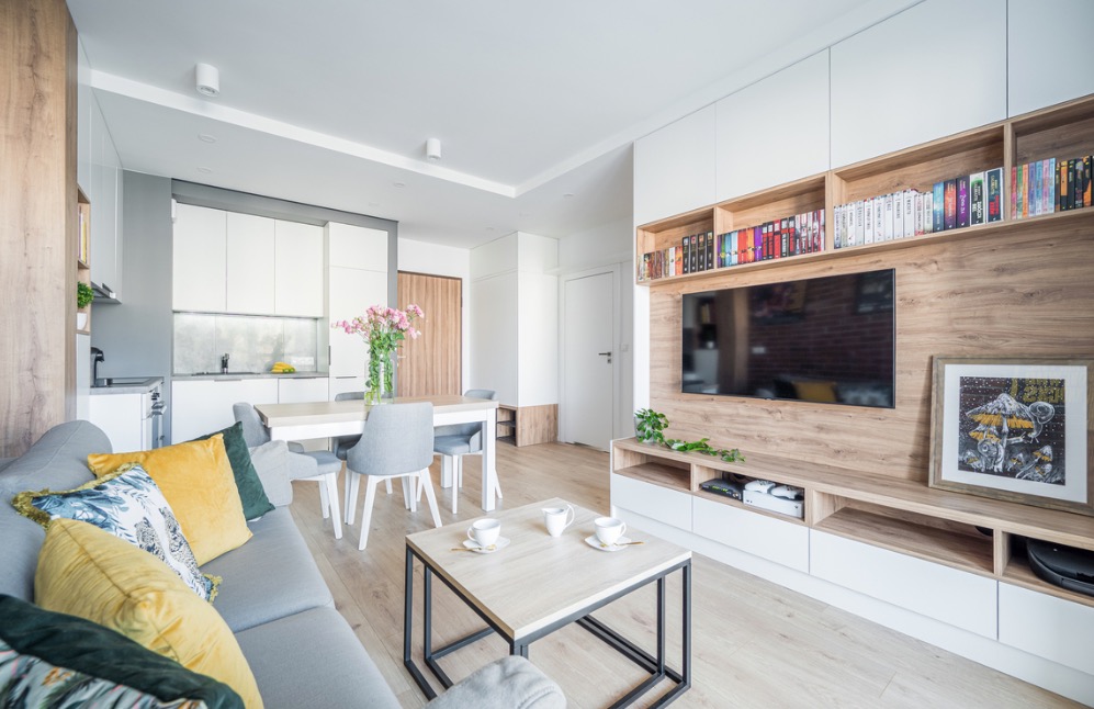 Jasne, przestronne wnętrze mieszkania z drewnianymi akcentami, szarą sofą z kolorowymi poduszkami, stołem jadalnym z kwiatami i ścianą telewizyjną z półkami na książki.