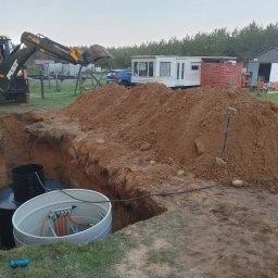 Rateko Robert Szczurowski - Żółta koparka MST przy wykopie, w którym umieszczono dwa zbiorniki, jeden czarny, drugi szary z instalacją, obok sterta ziemi, w tle przyczepa kempingowa i budynek gospodarczy.