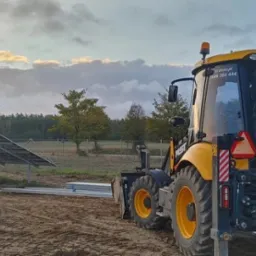 Żółto-czarna koparko-ładowarka na placu budowy instalacji fotowoltaicznej, widoczne panele słoneczne w tle, ziemia z wyraźnymi śladami opon, pochmurne niebo oświetlone promieniami zachodzącego słońca.