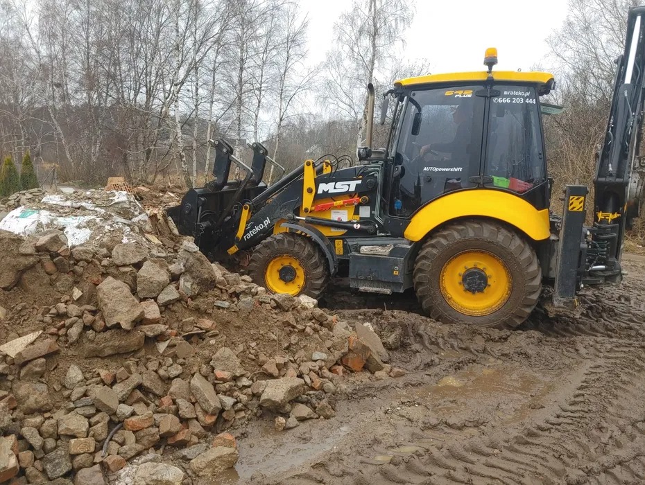Żółto-czarna koparko-ładowarka MST w błotnistym terenie, pracująca przy pryzmie gruzu i ziemi, z widocznym numerem telefonu na kabinie i drzewami w tle.