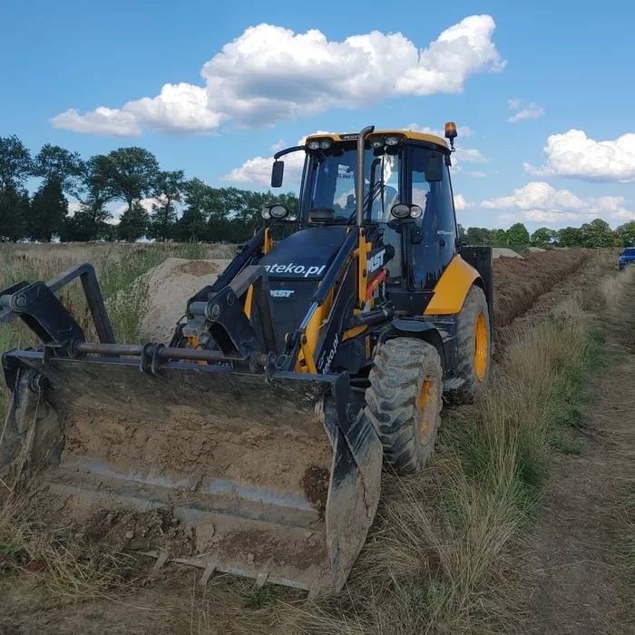 Żółto-czarna koparko-ładowarka z łyżką wypełnioną ziemią na tle wykopu w polu, błękitne niebo z obłokami, widoczny fragment lasu w oddali.