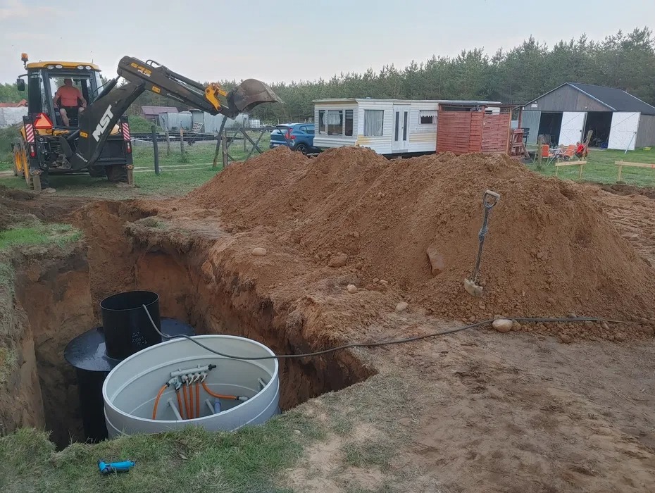 Żółta koparka MST przy wykopie, w którym umieszczono dwa zbiorniki, jeden czarny, drugi szary z instalacją, obok sterta ziemi, w tle przyczepa kempingowa i budynek gospodarczy.