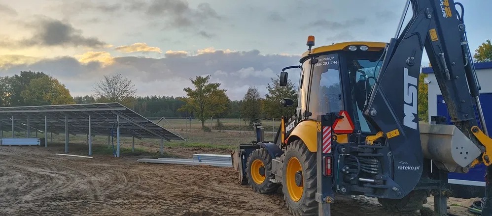 Żółto-czarna koparko-ładowarka na placu budowy instalacji fotowoltaicznej, widoczne panele słoneczne w tle, ziemia z wyraźnymi śladami opon, pochmurne niebo oświetlone promieniami zachodzącego słońca.