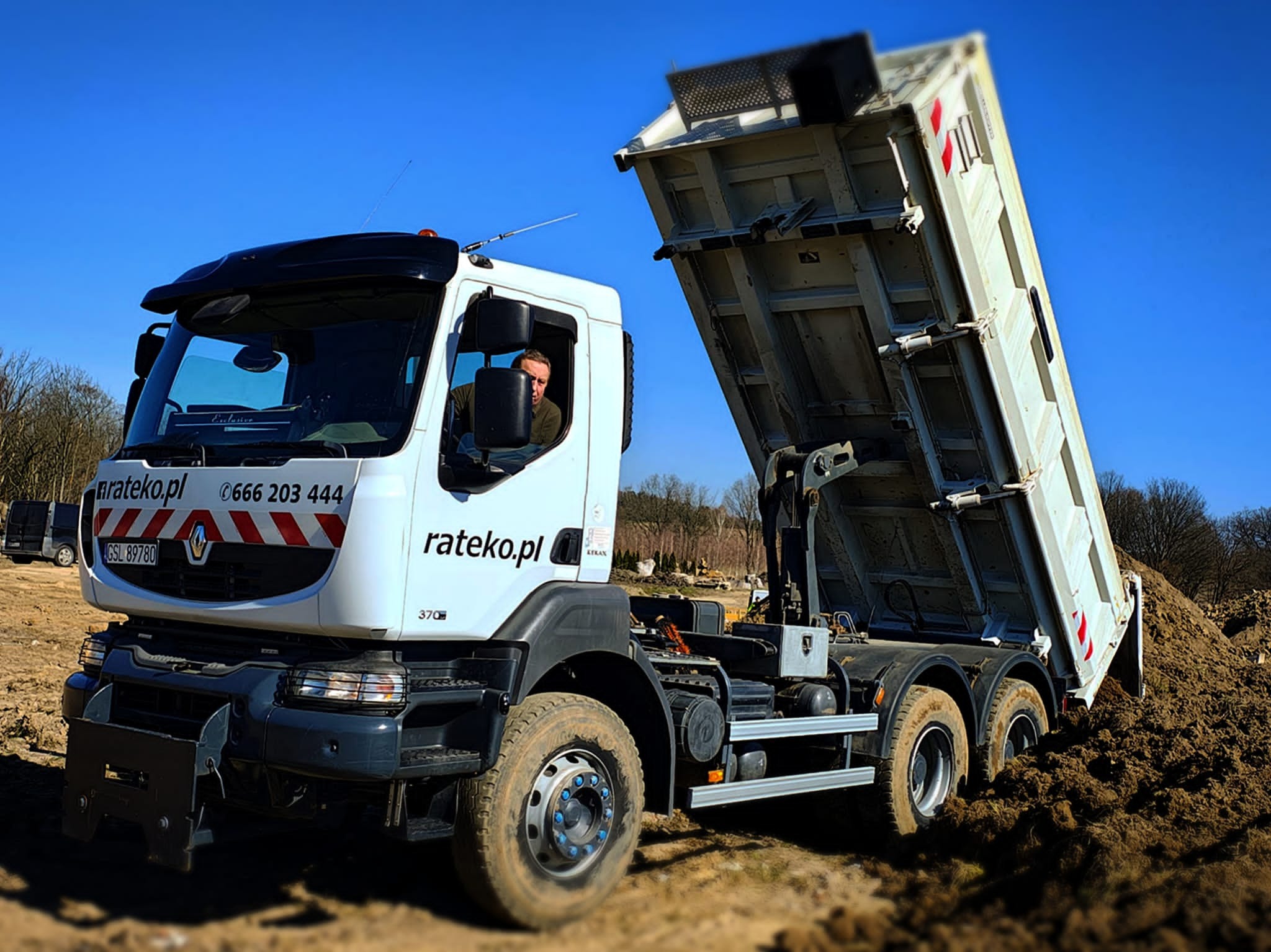 Wyładowujący ziemię biały samochód ciężarowy Renault z podniesioną skrzynią i numerem telefonu na kabinie, na tle błękitnego nieba i drzew.