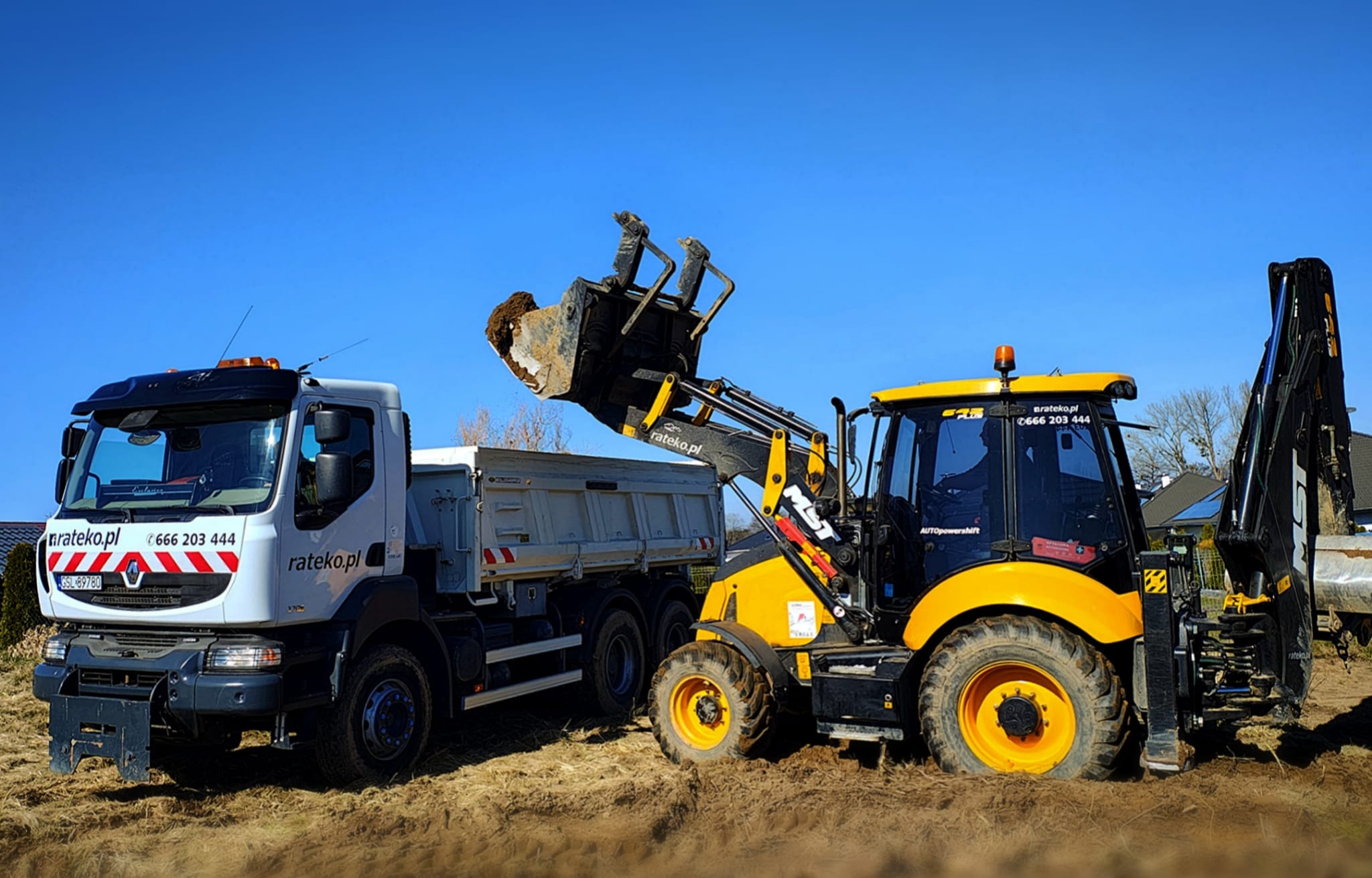 Żółto-czarna koparko-ładowarka wysypuje ziemię do białej wywrotki na tle błękitnego nieba. Widoczne numery kontaktowe i adresy stron internetowych na pojazdach.